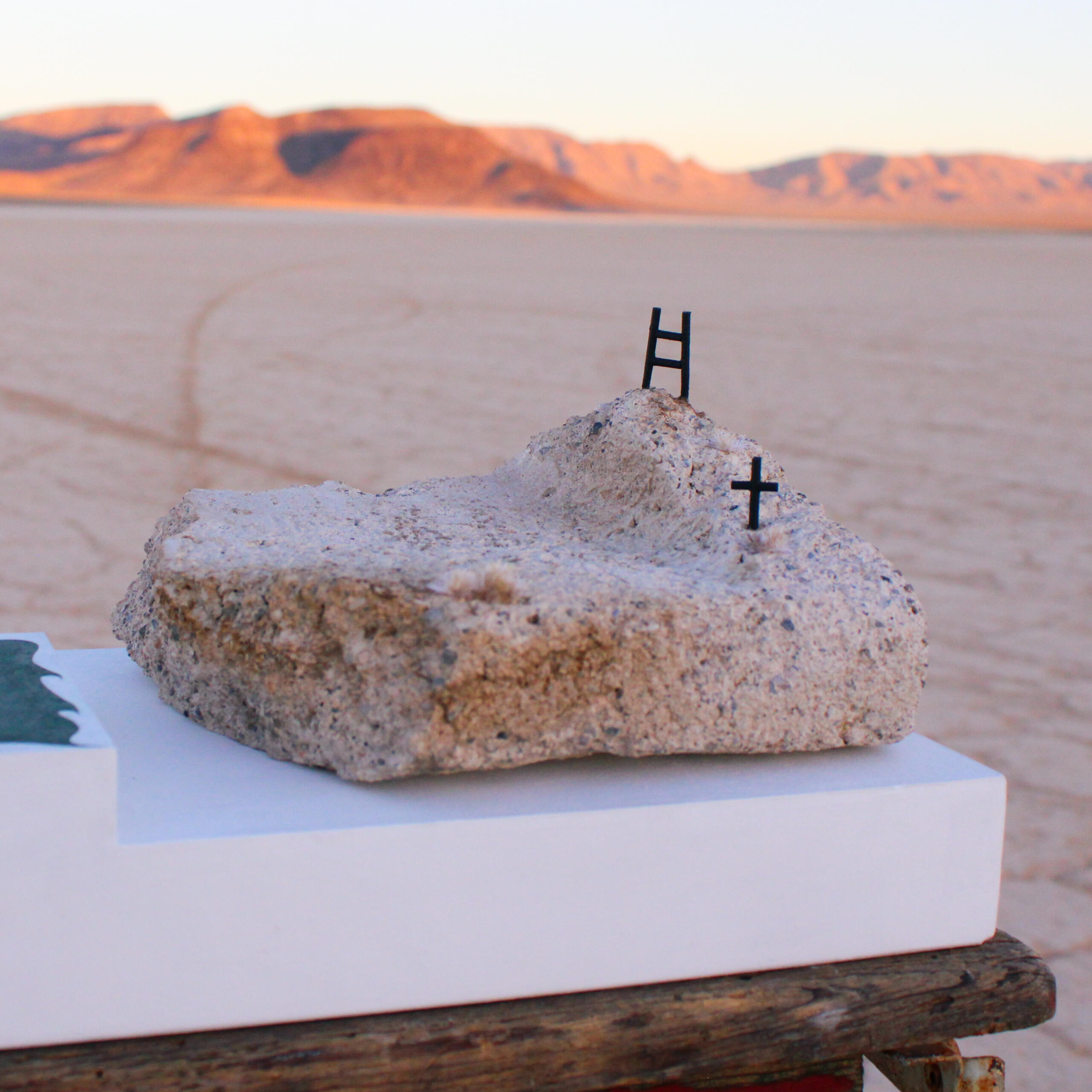Image from Stone Soup event from 2023 Desert Biennial Project. Features rock on white pedestal with miniature ladder on top of rock and miniature cross slightly lower. Artwork by Mary Sabo.