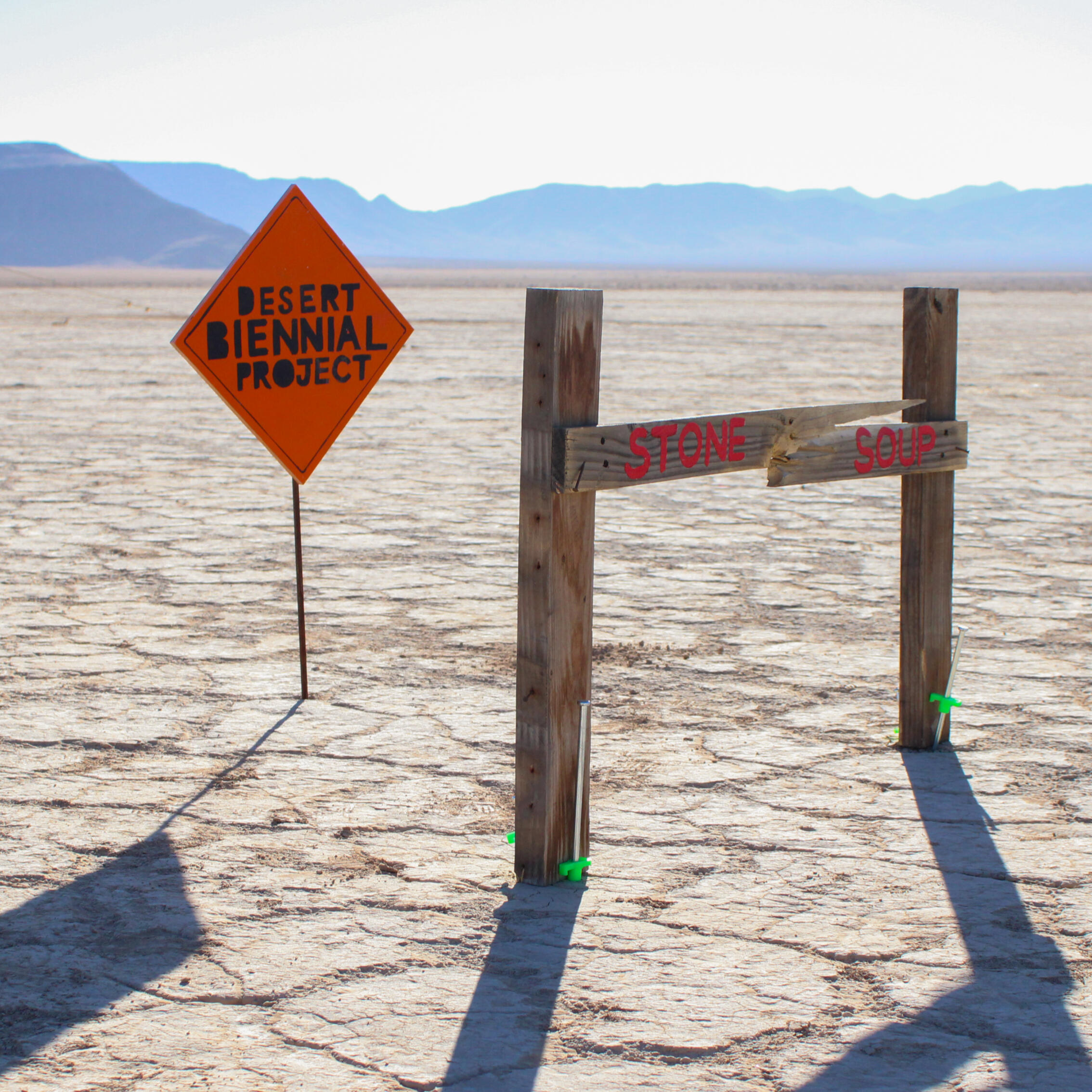 Image from Stone Soup event from 2023 Desert Biennial Project. Features bright orange diamond sign with black lettering spelling out Desert Biennial Project behind a sign made from dimensional lumber with Stone Soup painted in red on front of sign.